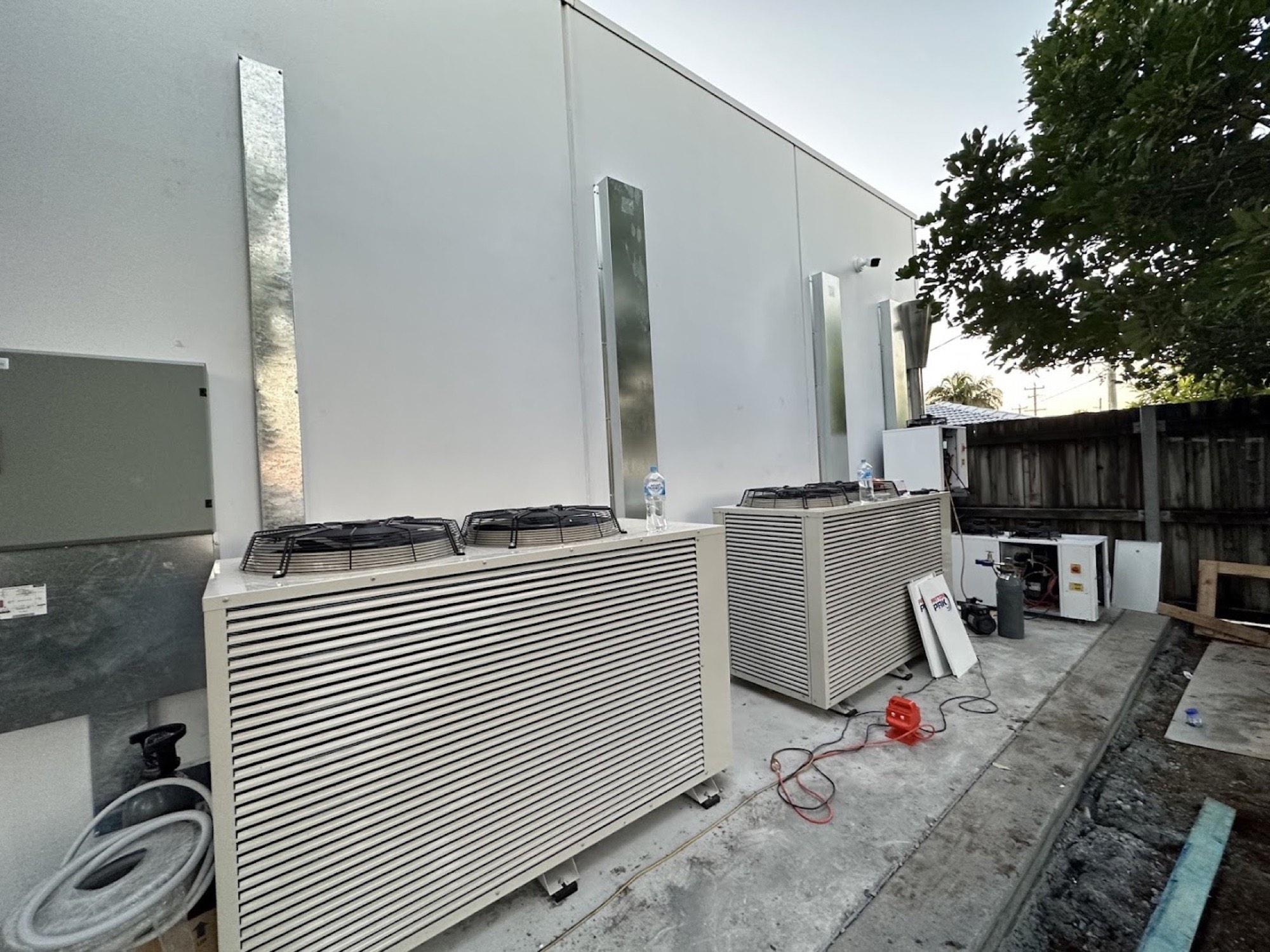 A row of large air conditioning condensers installed on a concrete plinth beside a building