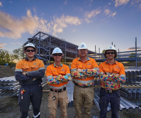 Four tradesmen in hi-vis standing in front of a steel building frame at dusk